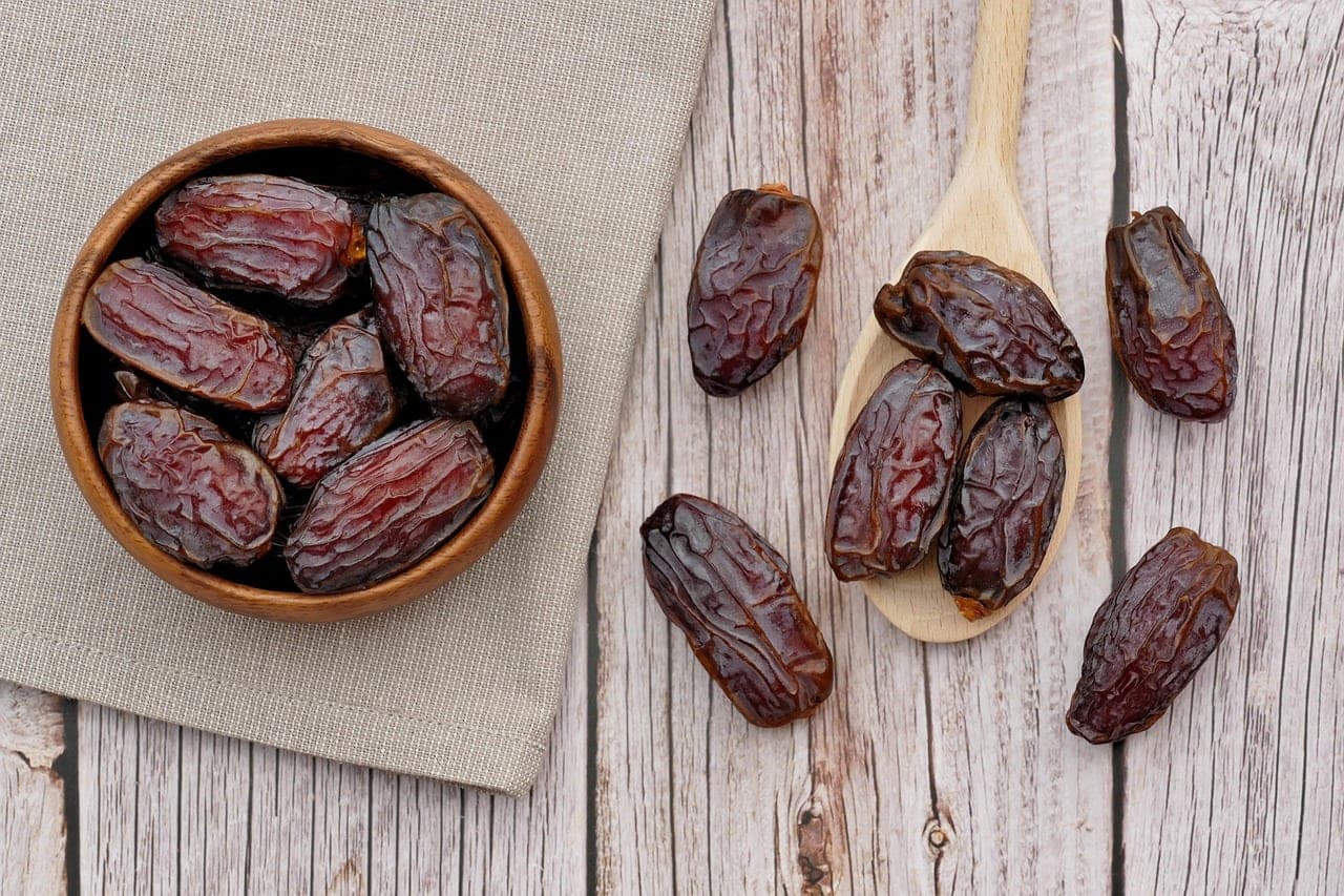 Brown dates in a wooden bowl and on a wooden spoon, set on a pale plank table.