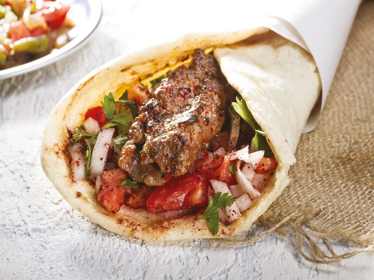 A grilled meat sandwich in flatbread with tomato, onion and parsley, served in its paper wrapper.