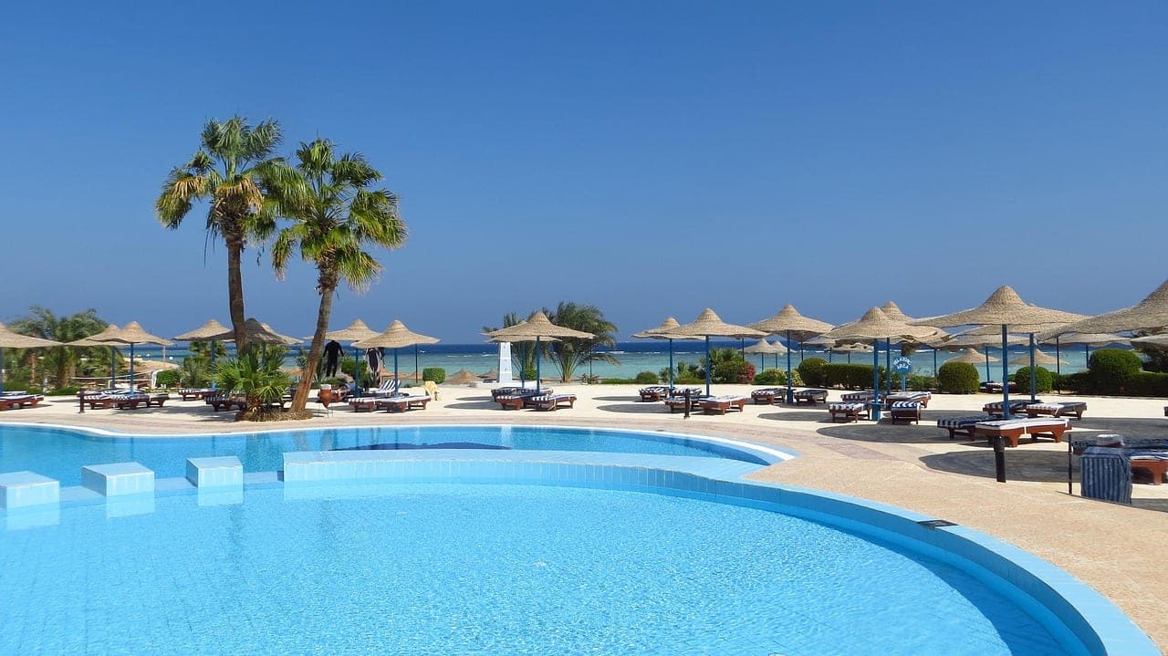 A blue pool in the foreground, a row of straw parasols, palm trees and the sea behind.