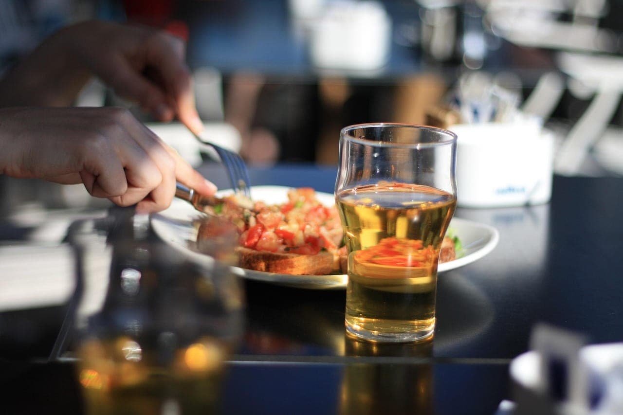 Two hands cut into a tomato-topped toast beside a glass of beer on a dark table.