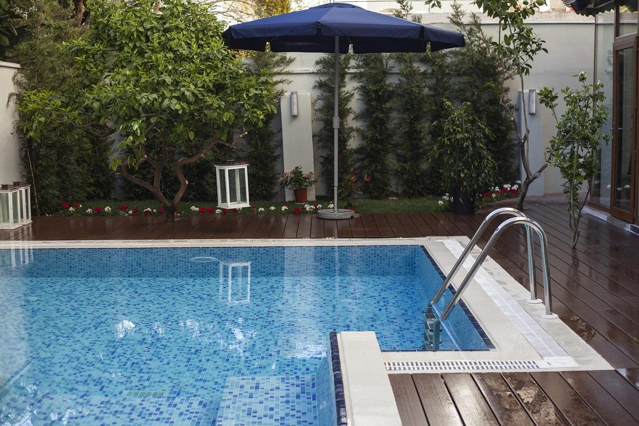 A square pool with blue mosaic tiling, an open parasol and a wooden deck, between a wall and trees.