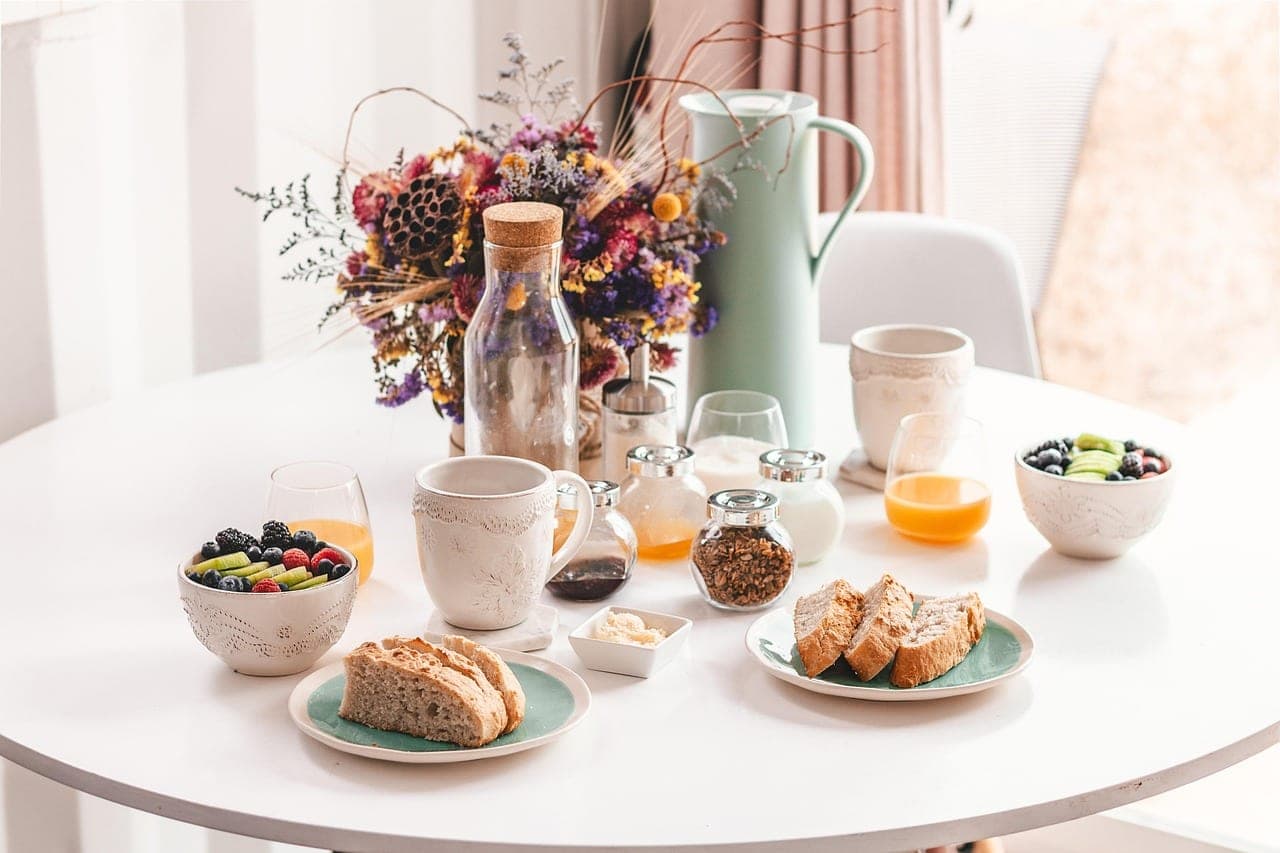 A brunch table set by a window: bread, fresh fruit, orange juice and coffee.