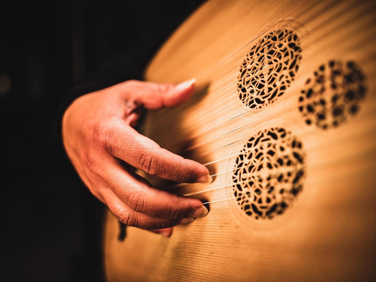 A hand plucks the strings of an oud, above the carved rosettes of its soundboard.