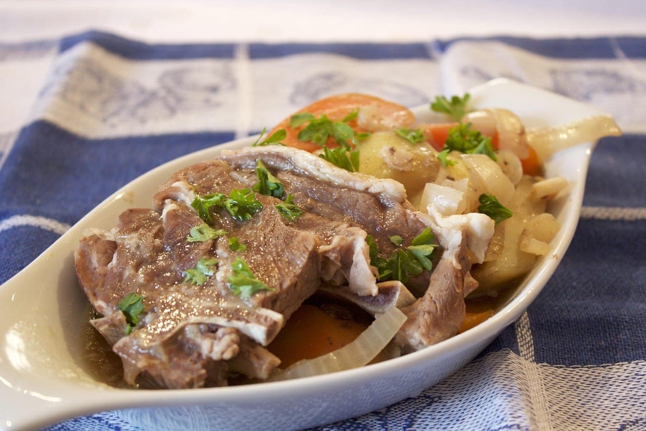 A piece of lamb slow-cooked with potatoes, carrots and onions in a white dish.