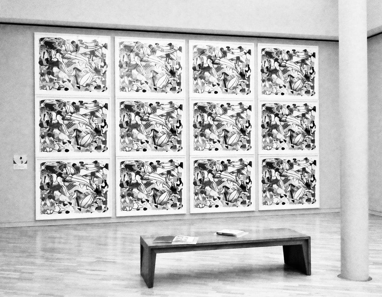 An exhibition wall: twelve framed prints and a bench, in black and white.