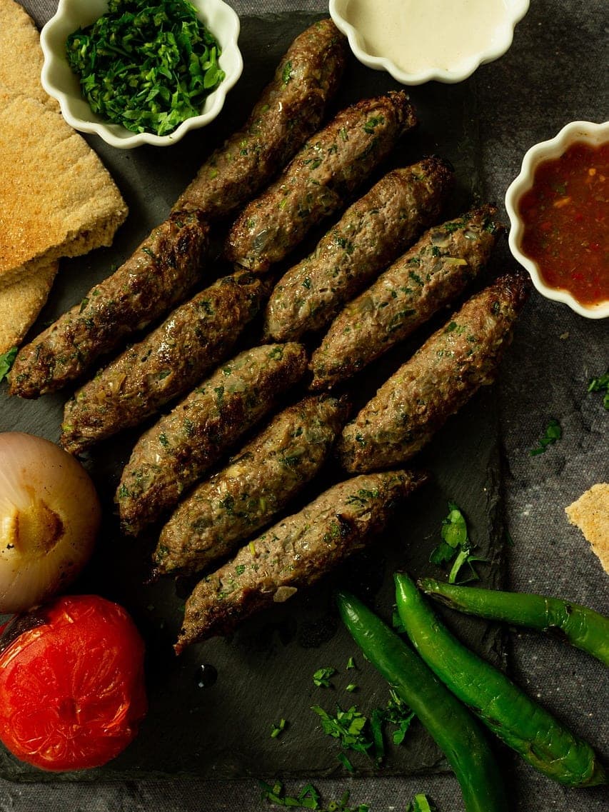 Rows of grilled kofta fingers on slate, with tahini, red sauce, grilled tomato and green chillies.
