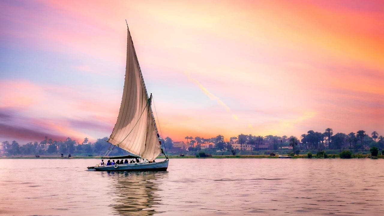 A white-sailed felucca crossing the river at sunset.