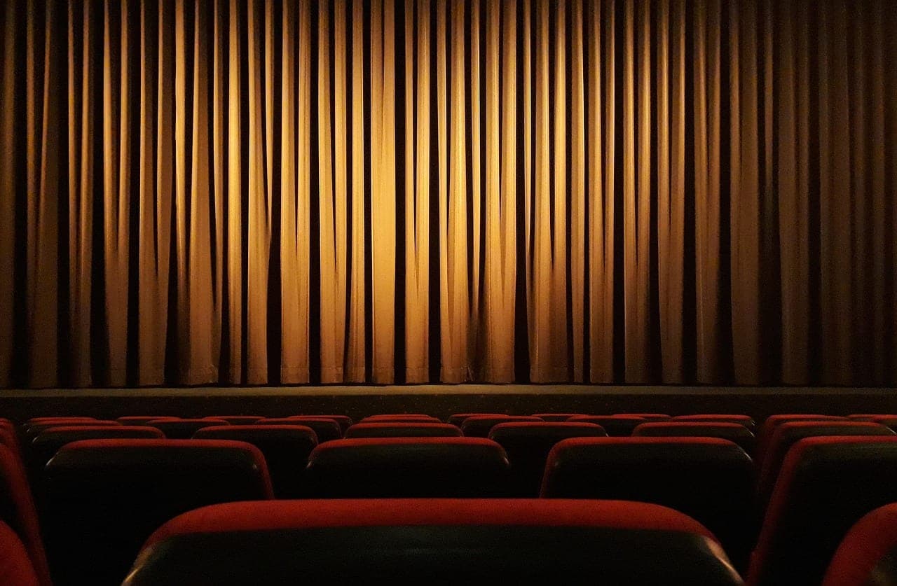 A large golden curtain lit from within, above the rows of empty red seats of a darkened hall.