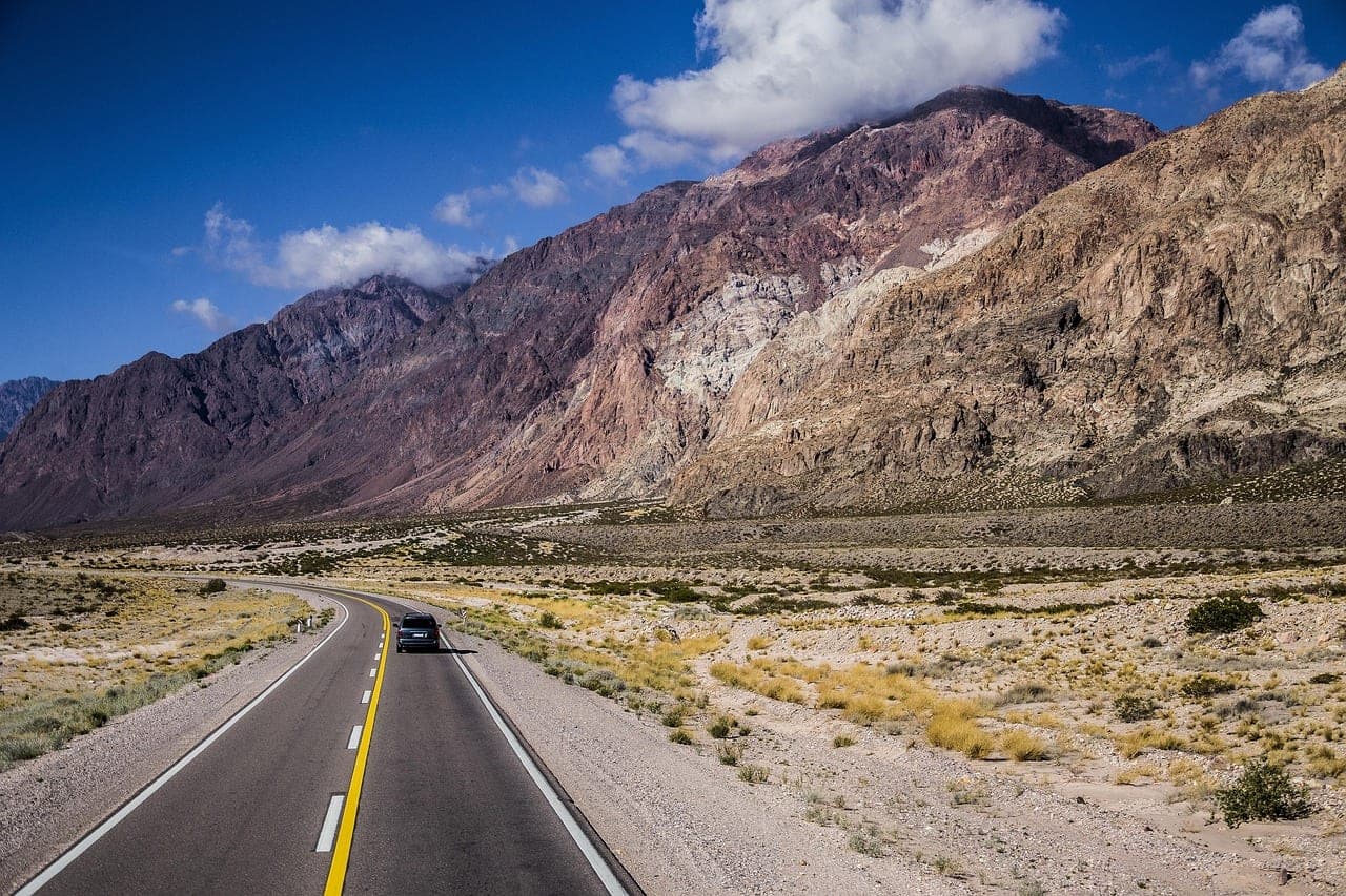 A desert road at the foot of a cliff, a lone car in the distance.