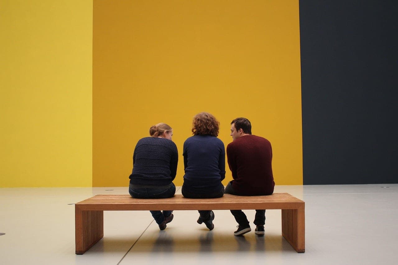 Three visitors sitting with their backs to us on a wooden bench, facing a large yellow wall in an exhibition room.