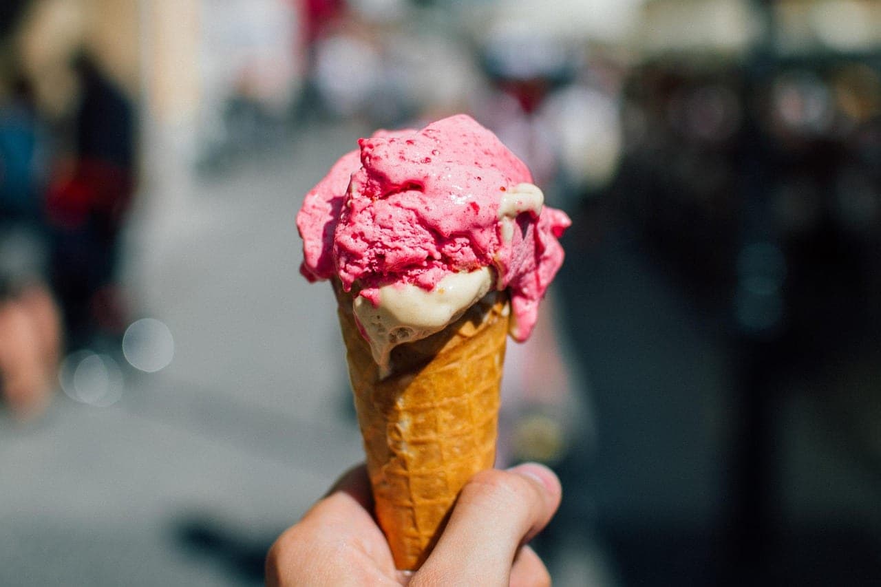 A hand holding an ice cream cone with a pink scoop and a cream scoop, out in the street.