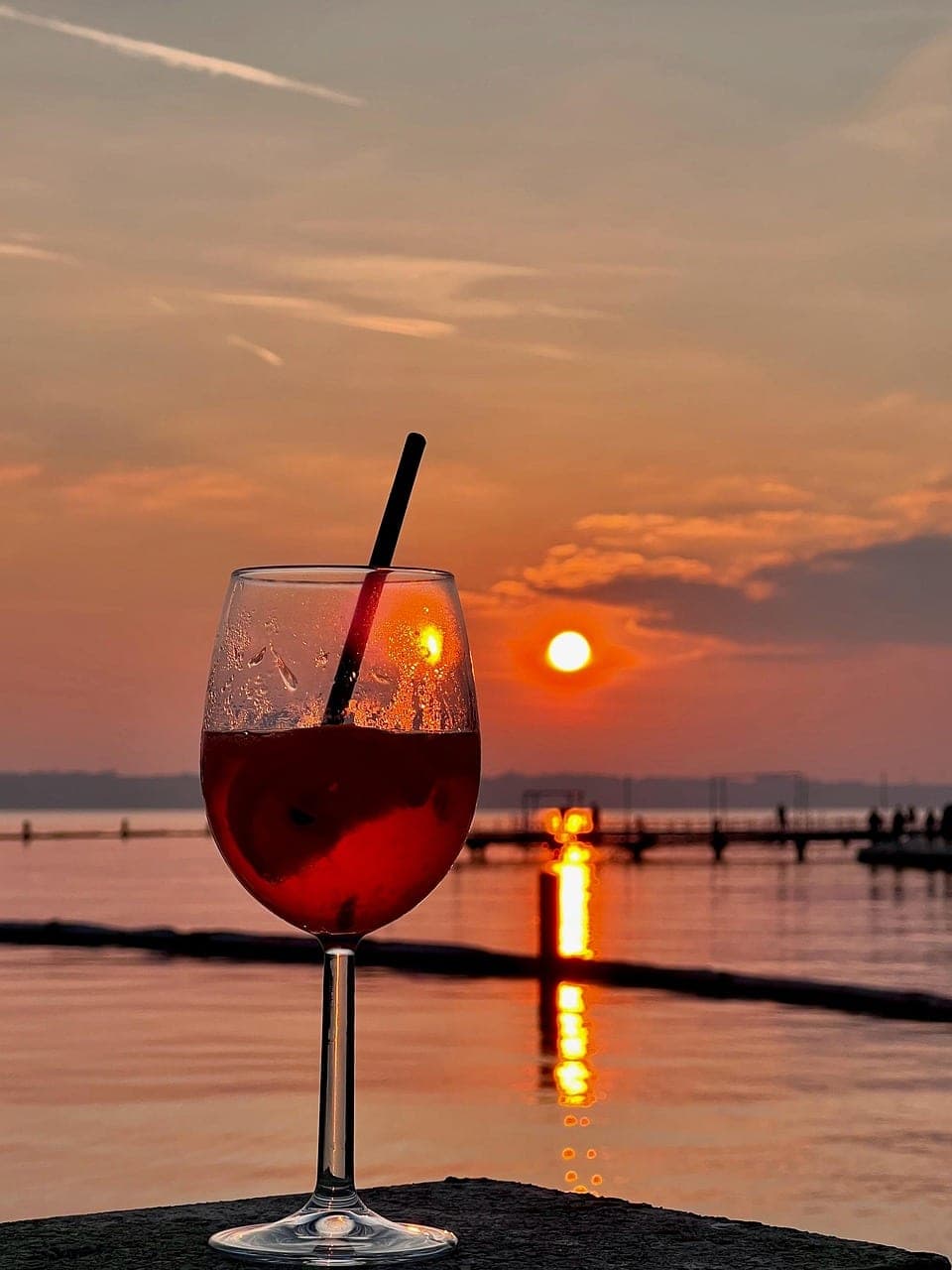 A red cocktail with a straw, set facing the sun as it drops over the water.