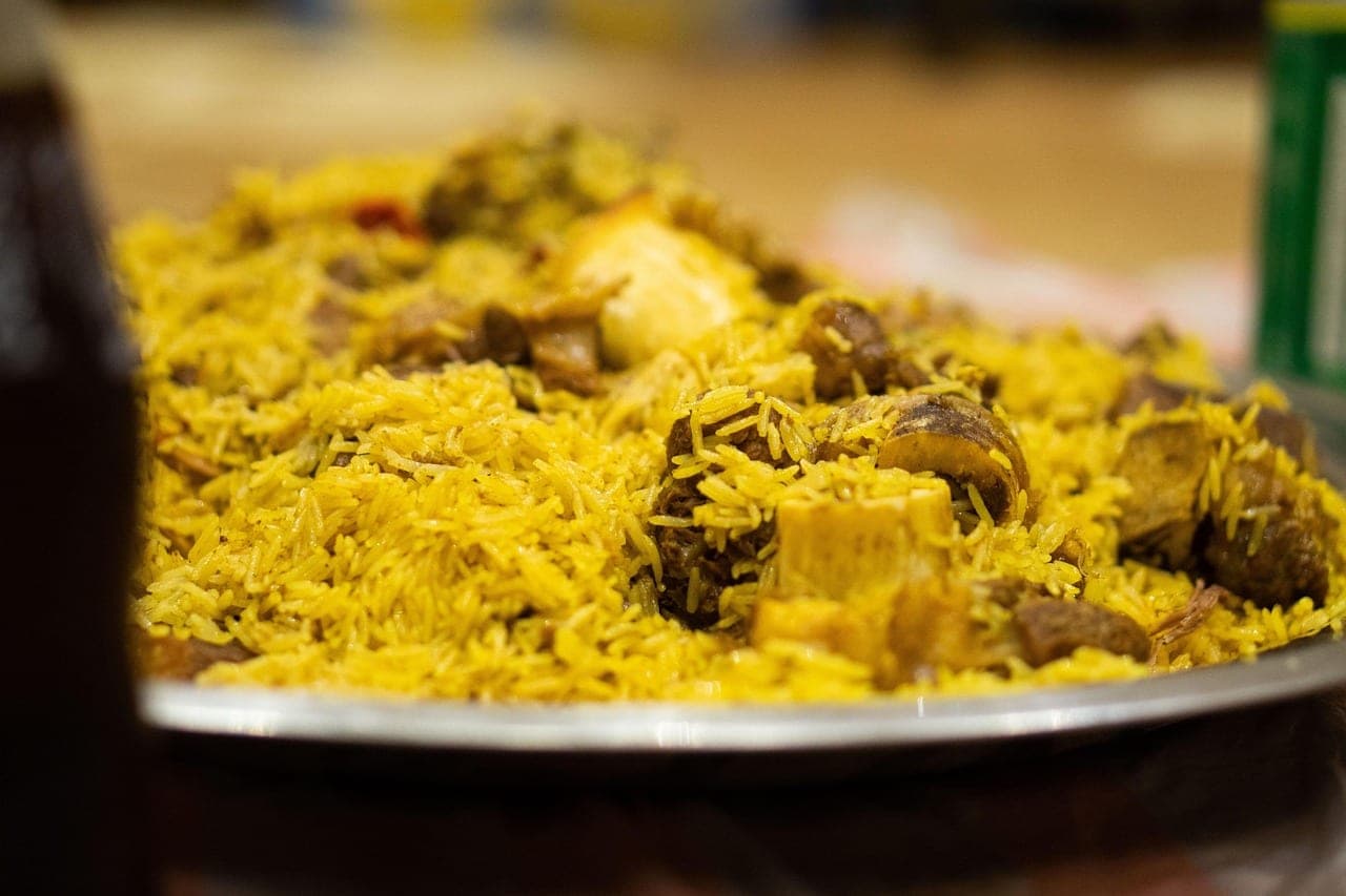 A large metal tray heaped with spiced yellow rice and pieces of meat, one with a lamb bone.