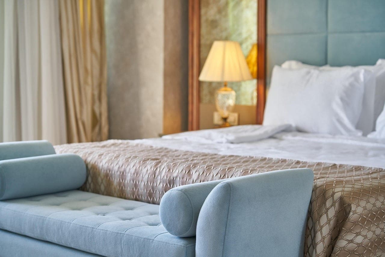 A hotel room, bed made and bedside lamp lit.