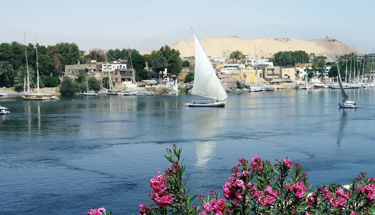 A felucca under sail on the Nile, before a village and a sand hill.