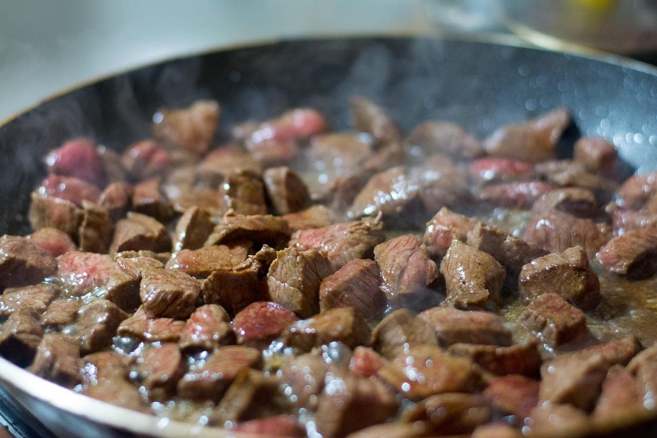 Chunks of meat sizzling in a large frying pan, steam rising.