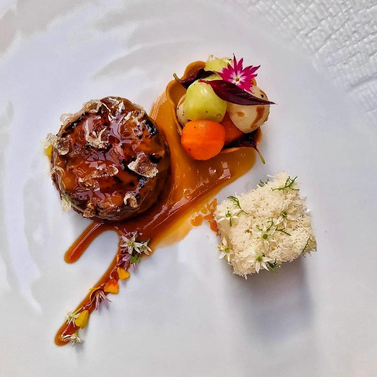 A chef-plated dish: a piece coated in amber sauce, glazed pearls and edible flowers.