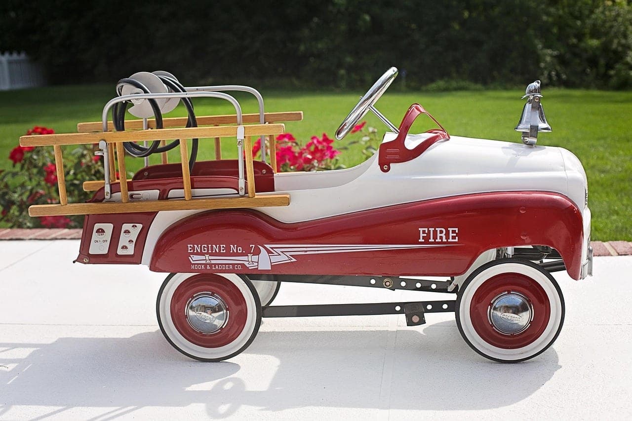 A child's pedal fire engine parked on a driveway in front of a lawn.
