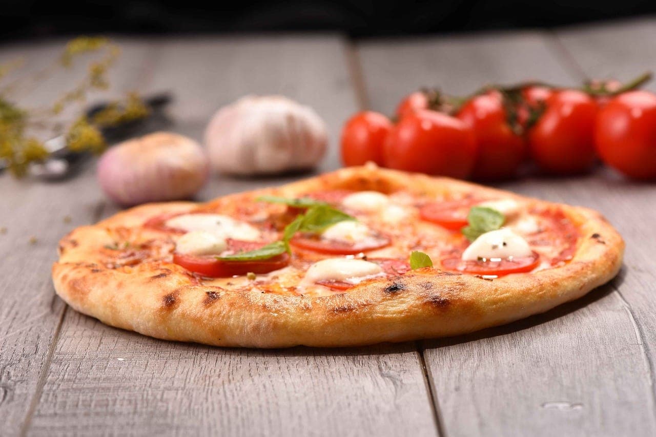 A margherita pizza on a wooden table, tomatoes and garlic beside it.