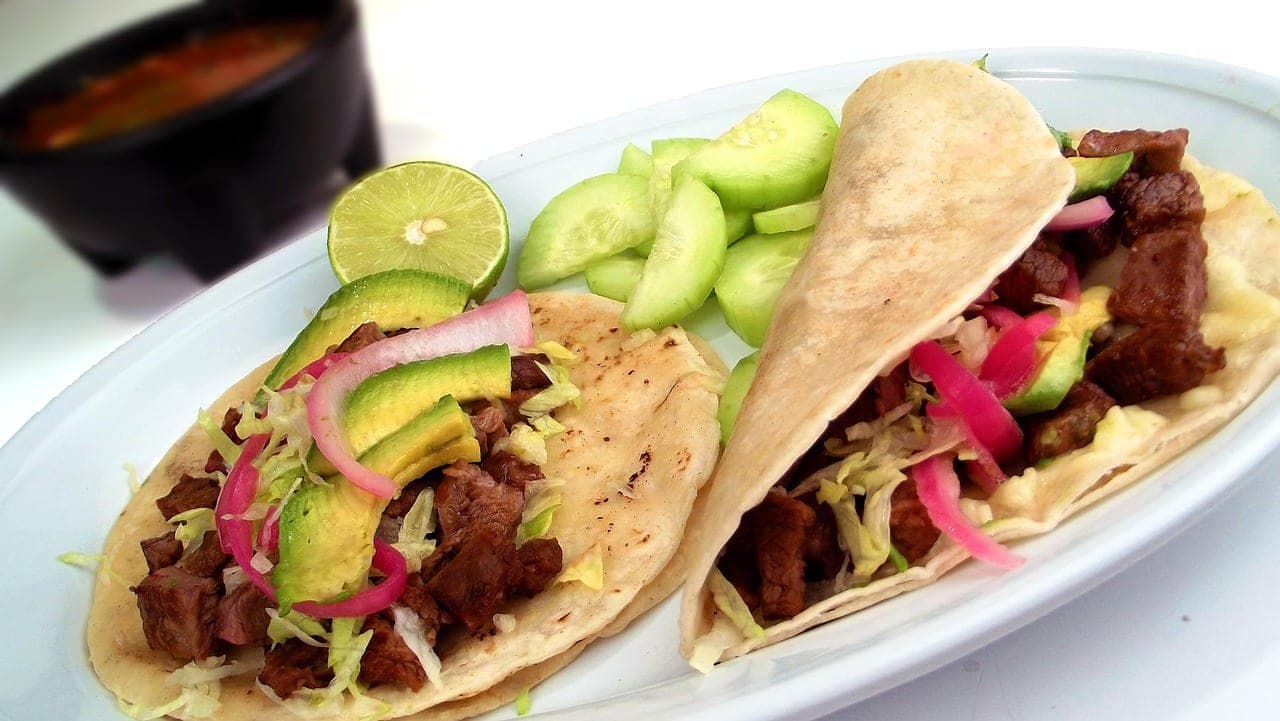 Two beef tacos topped with avocado, red onion and lime, on a white dish.