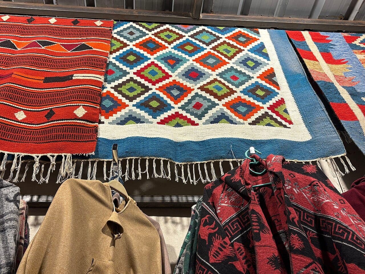 Woven rugs with geometric patterns hung above clothes in a craft shop.
