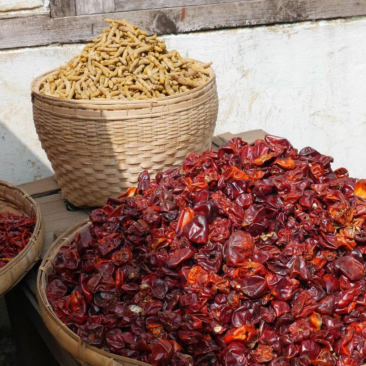 A mound of dried chillies in a woven tray, beside a basket of turmeric roots.
