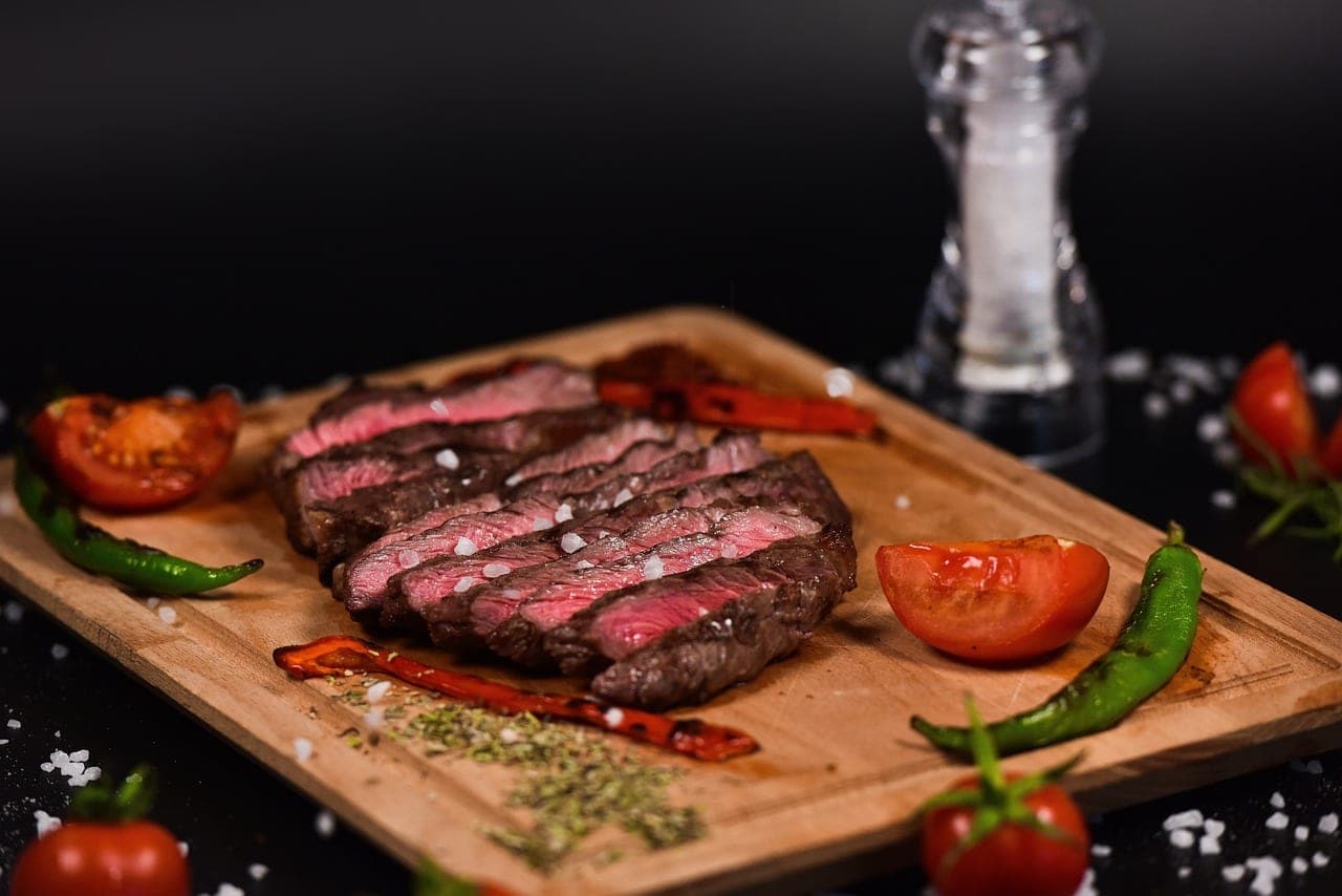 A sliced rare beef steak on a wooden board, with grilled tomato and peppers.