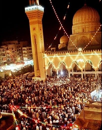 A mosque strung with lights at night, a dense crowd filling the square in front of it.