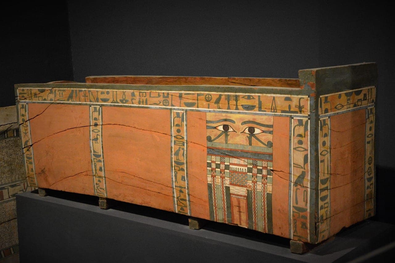A painted wooden sarcophagus, covered with hieroglyphs and two wedjat eyes, on its plinth in a museum gallery.