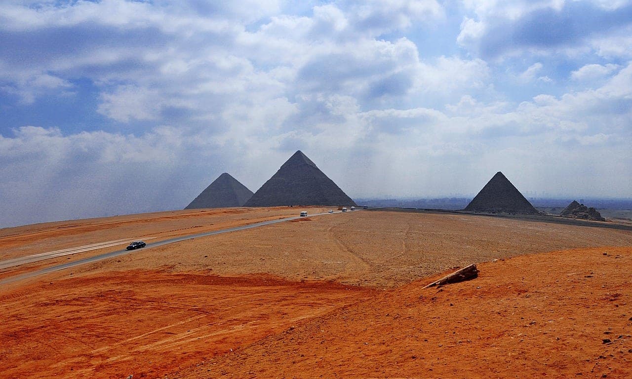 A road crosses the desert plateau, a few vehicles in front of the three pyramids.