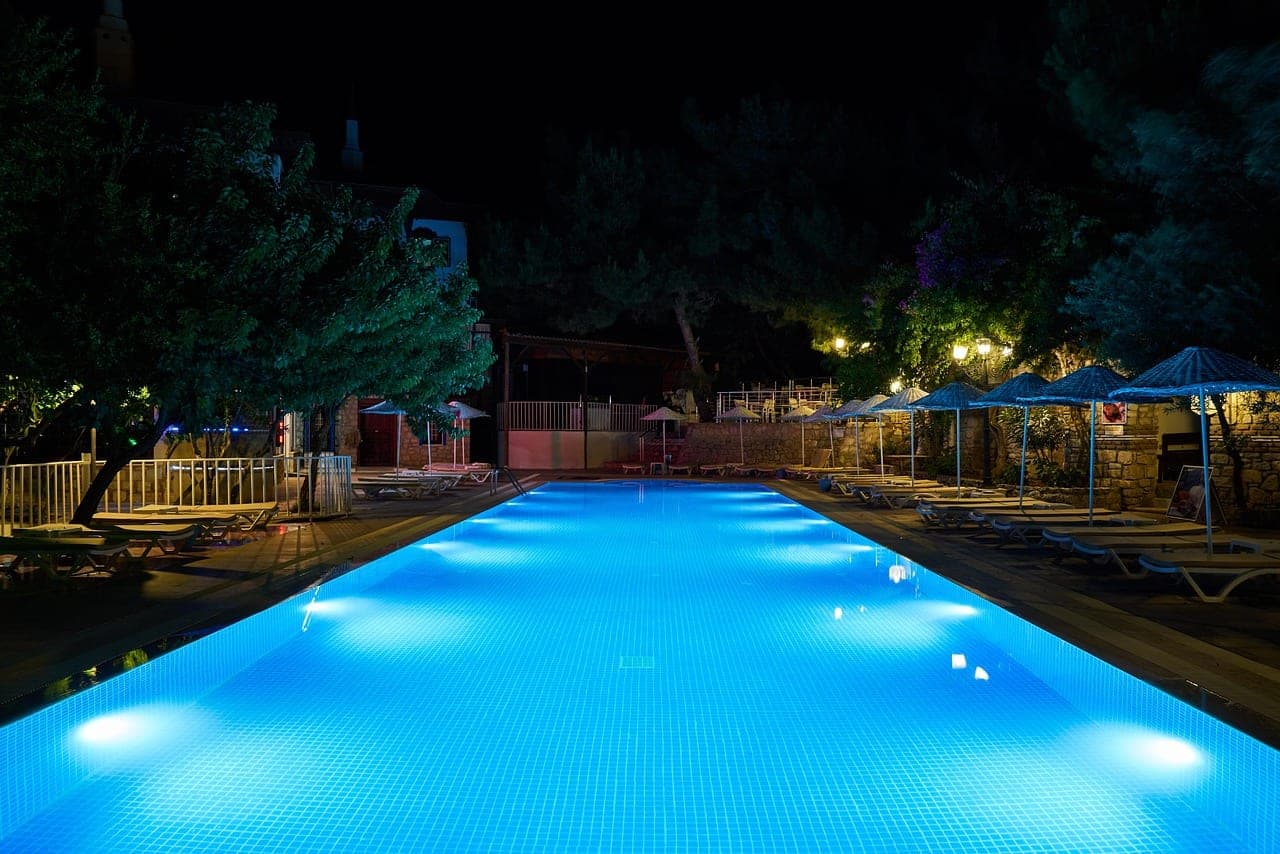 A pool lit blue at night, edged with empty loungers and straw parasols.