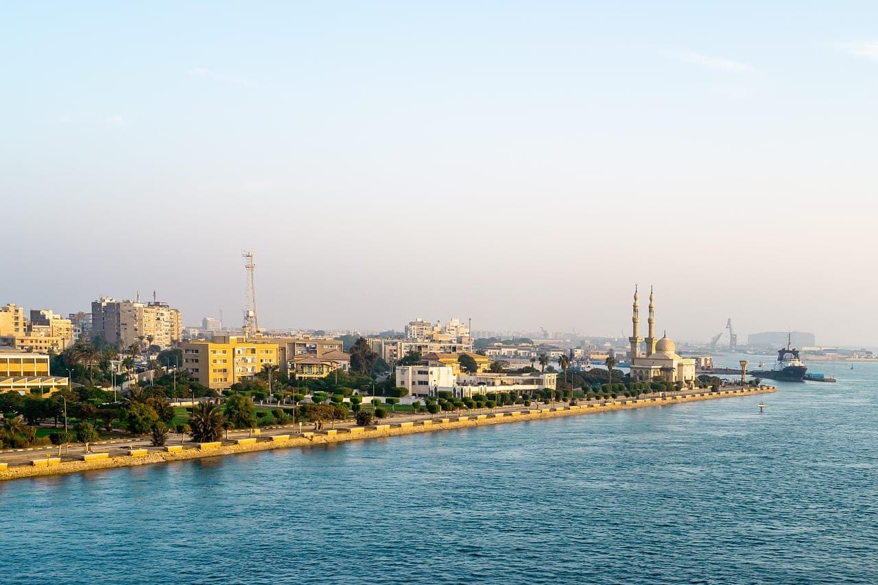 A palm-lined corniche along the Suez Canal, a mosque at the water's edge and a tug moored at the quay.