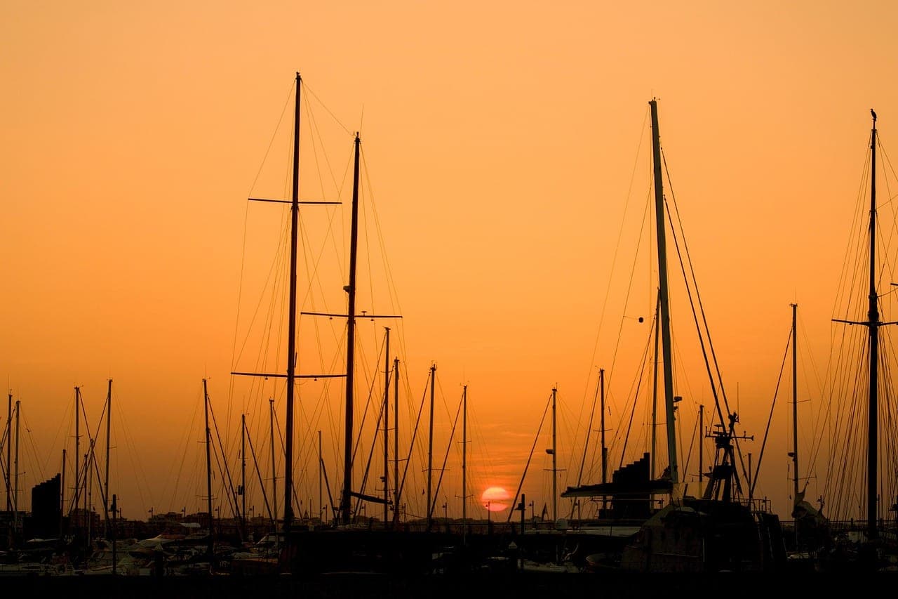 Sailboat masts silhouetted against an orange sky as the sun sets over a marina.