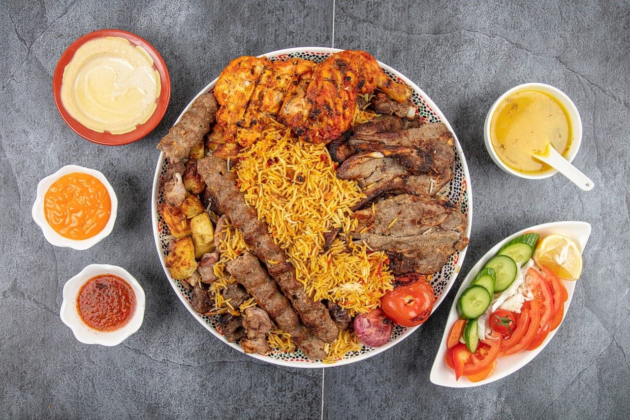 A large platter of yellow rice topped with kofta skewers, chicken and grilled meats, ringed with small bowls of sauces.