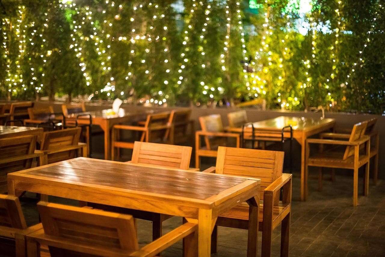 An open-air wooden terrace in the evening, under a curtain of fairy lights and foliage.