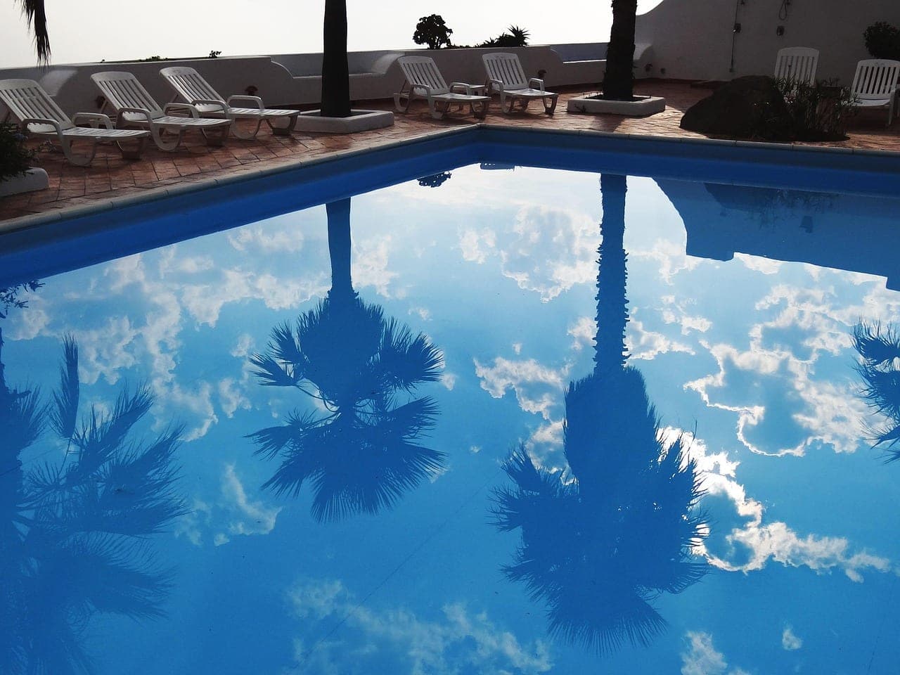 Palms and clouds mirrored in the blue water of a pool, white loungers along the edge.