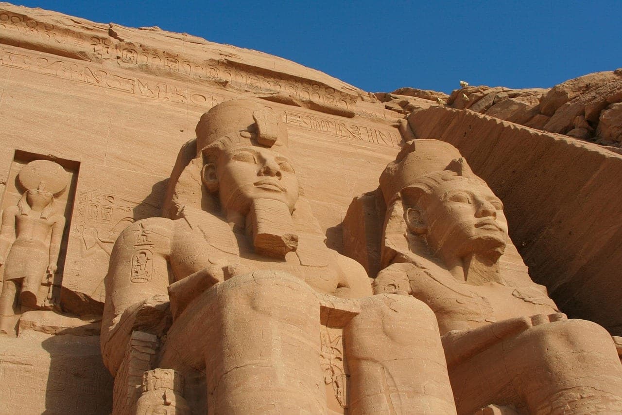 Two stone colossi carved into the cliff face, under a blue sky.
