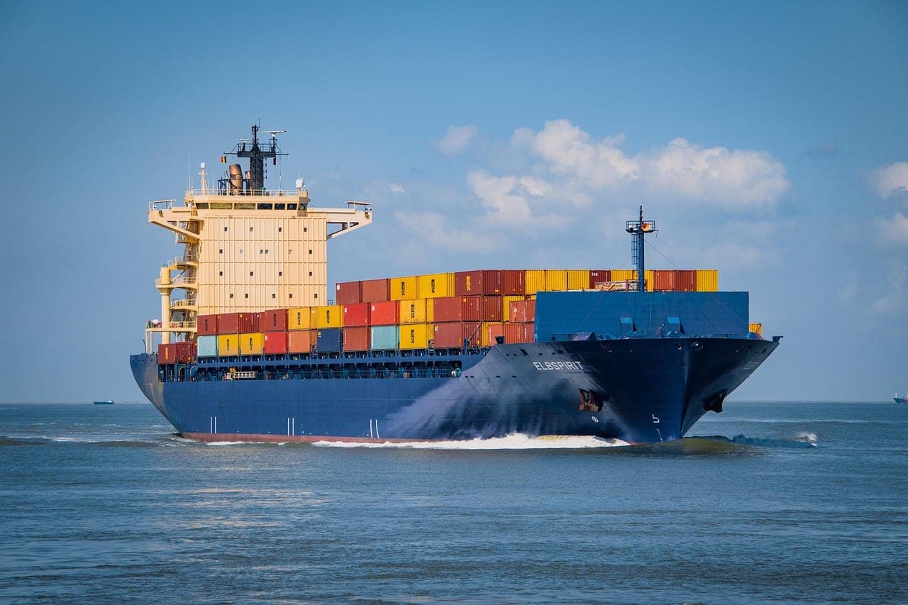 A loaded container ship seen head-on, cutting through open water.