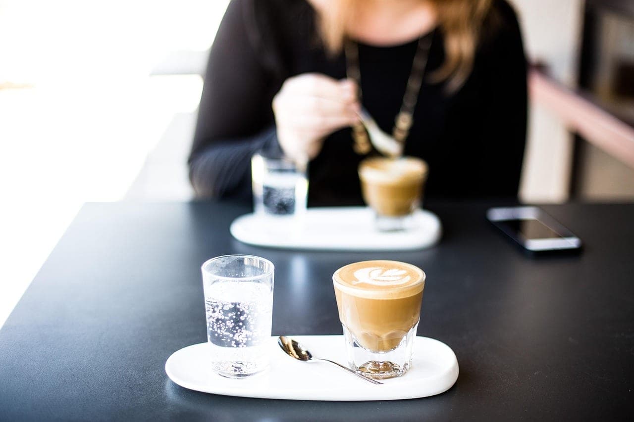 Two milky coffees in glasses, on white trays at a café table.