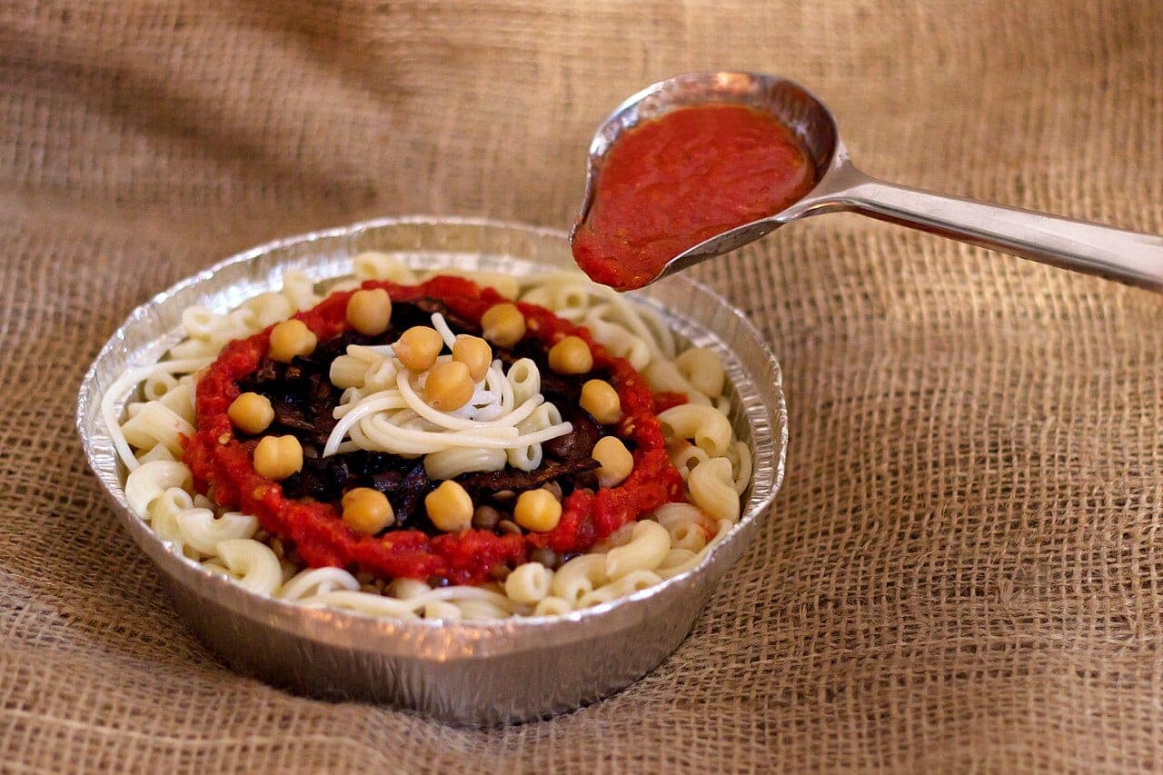 A foil tray of pasta, rice and chickpeas, topped with tomato sauce ladled over it.