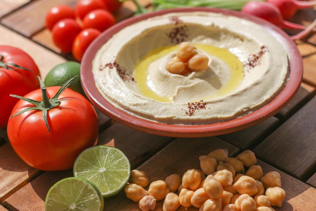 A dish of hummus with olive oil, ringed by tomatoes and limes, on a wooden table.