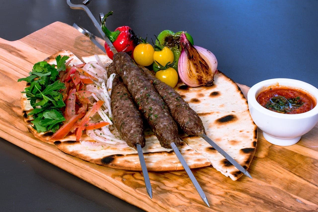 Three kofta skewers on flatbread with grilled vegetables, tomato salad and a red sauce.