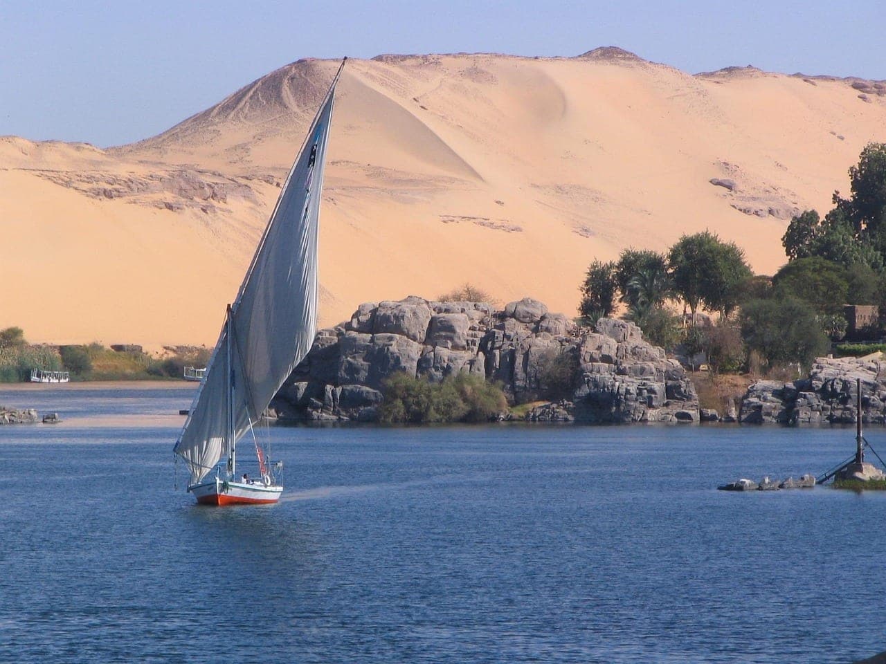 A white-sailed felucca on the Nile, in front of granite rocks and a tall sand dune.