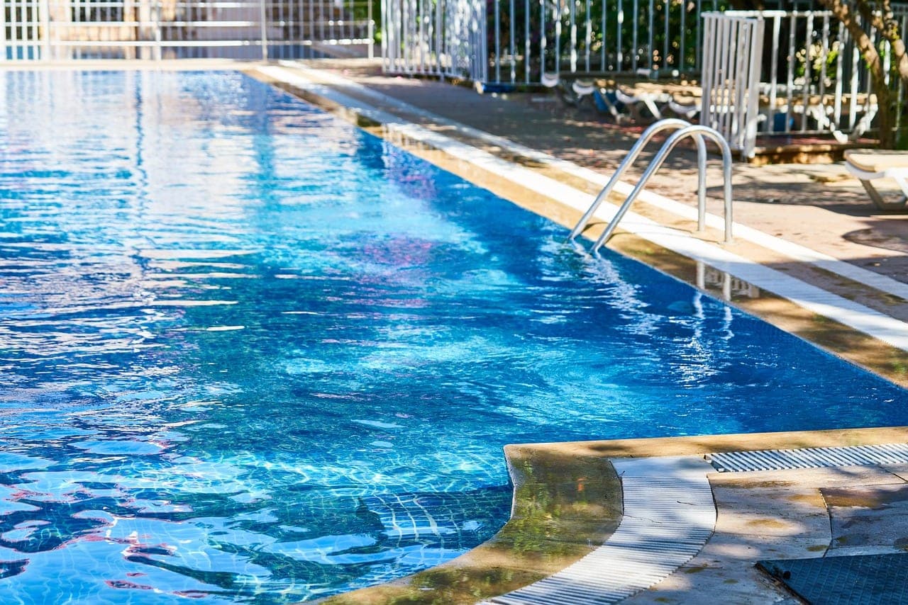 The edge of a pool in full sun, a metal ladder, empty loungers behind a railing.