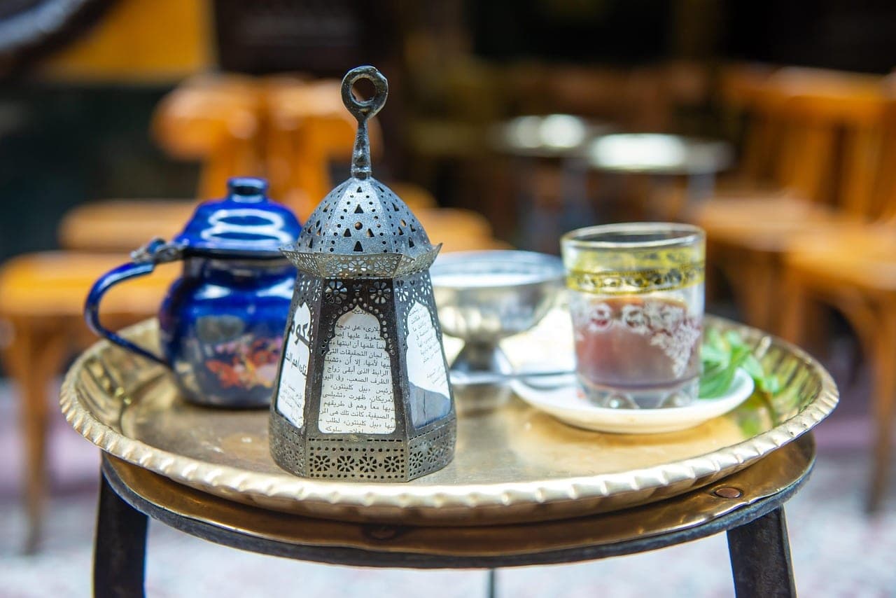 A pierced lantern, a teapot and a glass of mint tea on a brass tray, among wooden café chairs.